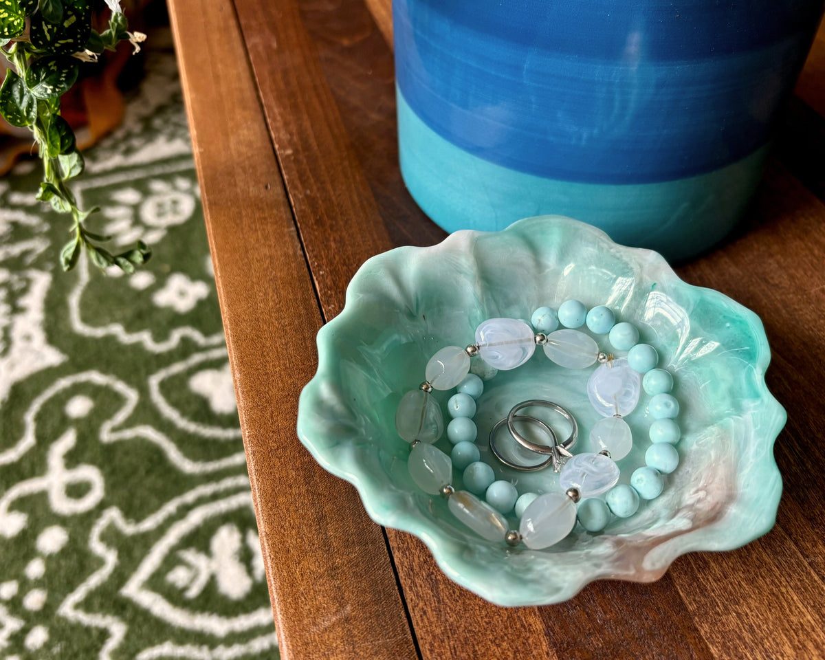 A resin flower-shaped jewelry tray in a light teal and copper pattern, holding a beaded bracelet and two rings, set on a wooden table next to a blue pot.