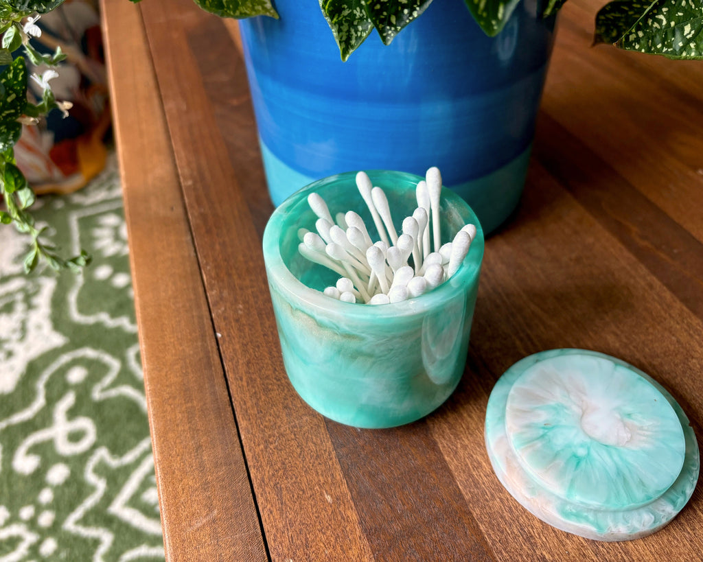 A small round turquoise resin container with a matching lid sits on a wooden table, filled with white cotton swabs. A larger blue planter and green plant are blurred in the background.