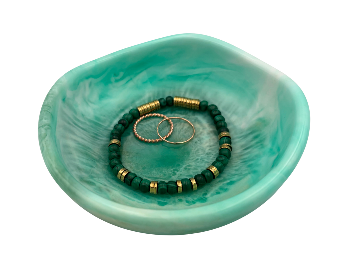 A small marbled turquoise-green resin dish holding two beaded bracelets and two gold rings, isolated on a white background.