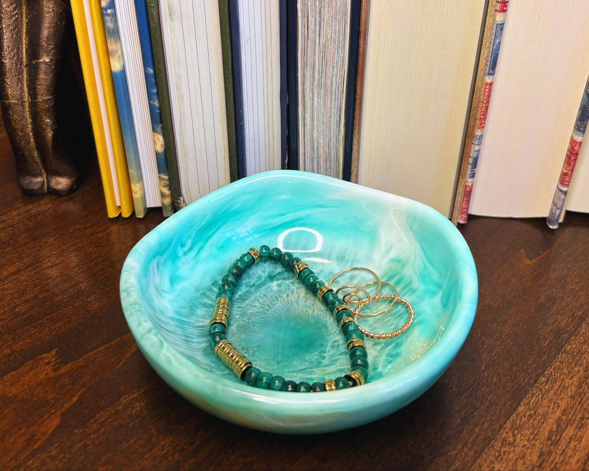 A small turquoise and white marbled resin bowl sits on a dark wooden table, styled with a green beaded bracelet and gold rings in front of a row of standing books.
