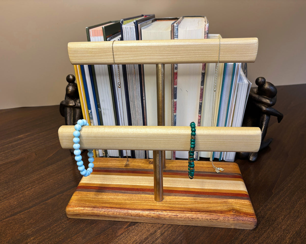 A wooden jewelry display stand with two horizontal bars holding light natural wood dowels, one decorated with a light blue beaded bracelet and the other with a dark green beaded bracelet. The stand features a striped wood base and stands in front of a stack of books on a dark wooden table.