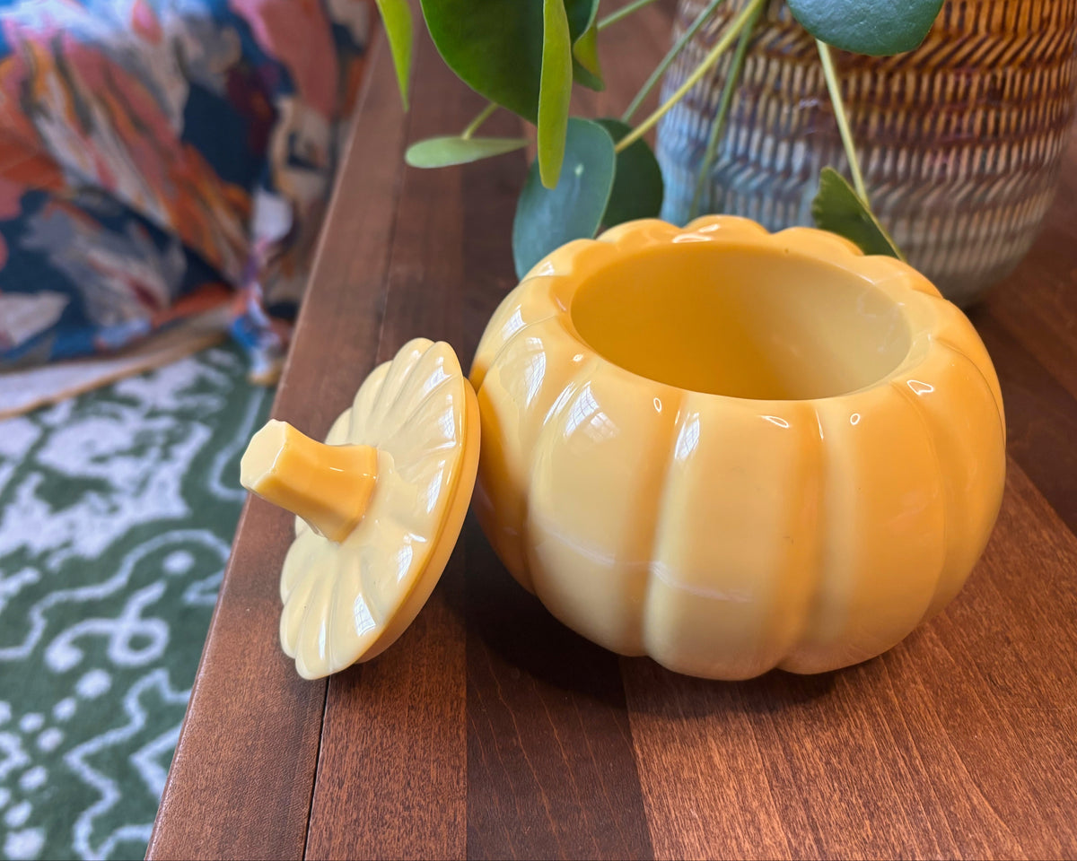 A glossy yellow resin pumpkin-shaped container with its lid leaning beside it on a wooden table, with a potted plant and patterned fabric in the background.