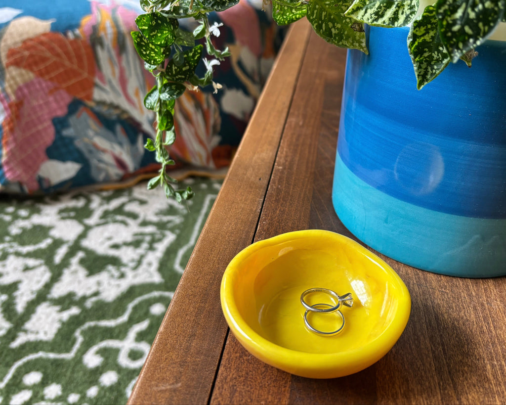 A small, shallow, bright yellow resin ring dish holding two silver rings, placed on a wooden table next to a blue and green glazed ceramic planter with a leafy plant.